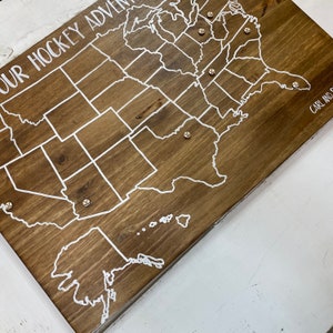 Puede incluir: Un mapa de madera de los Estados Unidos con contornos blancos de los estados y una inscripci&oacute;n blanca que dice "Our Hockey Adventure". Hay peque&ntilde;as tachuelas plateadas colocadas en el mapa.