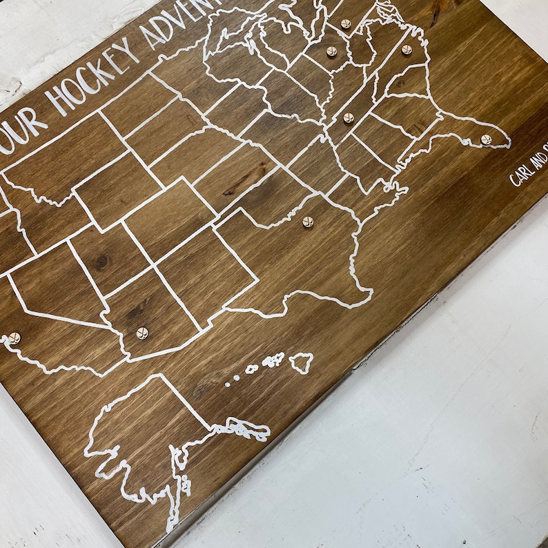 Puede incluir: Un mapa de madera de los Estados Unidos con contornos blancos de los estados y una inscripci&oacute;n blanca que dice "Our Hockey Adventure". Hay peque&ntilde;as tachuelas plateadas colocadas en el mapa.