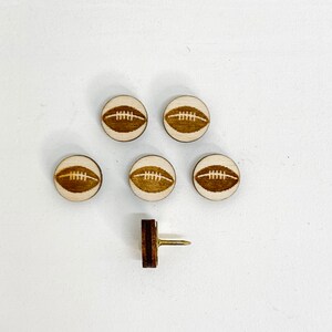 Puede incluir: Seis chinchetas de madera con un dise&ntilde;o de bal&oacute;n de f&uacute;tbol y una chincheta marr&oacute;n con una chincheta de metal dorado.