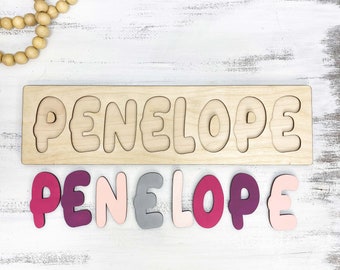 May include: Wooden puzzle spelling "PENELOPE" with colorful letters in shades of pink, purple, and gray. The puzzle is on a white wooden surface, with a wooden bead garland in the upper left corner. A fun educational toy.