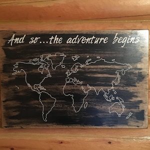 May include: A black wooden sign with a white outline of a world map and the text "And so...the adventure begins".