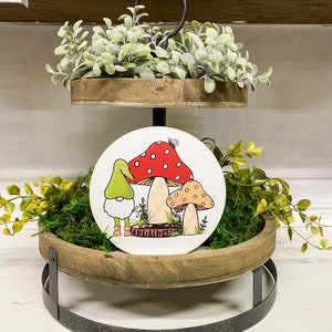 May include: A two-tiered wooden tray with greenery and a round wooden sign featuring a gnome and mushrooms. The sign has a red and white mushroom, a tan mushroom with orange spots, and a gnome wearing a green hat.