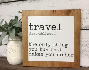 May include: A wooden sign with a white background and black text that reads "travel [trav-uhl] noun the only thing you buy that makes you richer".