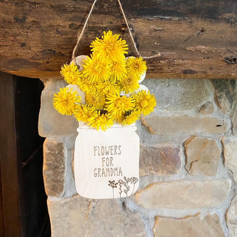 May include: A white wooden cutout of a mason jar with yellow dandelion flowers inside. The jar has the text "Flowers for Grandma" written on it.