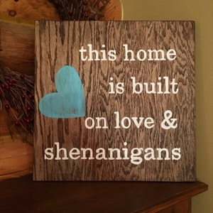 May include: A wooden sign with the text "this home is built on love & shenanigans" in white paint. A blue heart is painted on the left side of the sign.