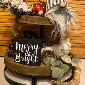 May include: A two-tier wooden tray with a black metal stand. The top tier has a red and white polka dot ornament, a gnome with a black and white striped hat, and a green and white sprig. The bottom tier has a pine cone, a wooden sign that says "Merry & Bright", a small white and black plaid fabric, a brown woven ball, and a sprig of green leaves.