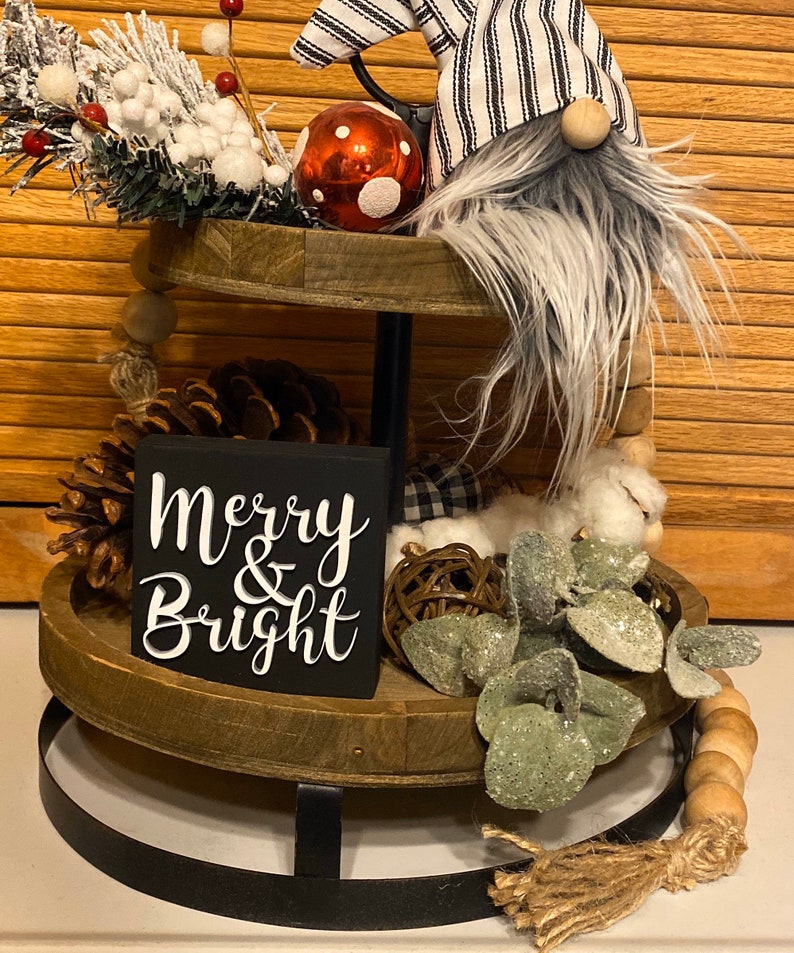 May include: A two-tier wooden tray with a black metal stand. The top tier has a red and white polka dot ornament, a gnome with a black and white striped hat, and a green and white sprig. The bottom tier has a pine cone, a wooden sign that says "Merry & Bright", a small white and black plaid fabric, a brown woven ball, and a sprig of green leaves.