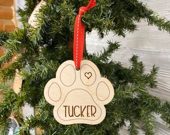 May include: A wooden paw print ornament with the name "TUCKER" engraved on it. The ornament is hanging from a red ribbon with white stitching and has a small heart cutout in the center.