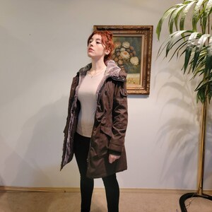 May include: A brown winter coat with a faux fur-lined hood and interior. The coat has multiple pockets and a cinched waist. The person in the image is wearing a light-colored top and black pants.