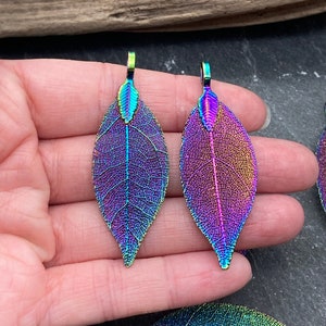 May include: Two iridescent leaf pendants with a rainbow color scheme of purple, blue, and green. The leaf pendants have a detailed vein structure and a small loop at the top for hanging. The leaves are approximately 3 inches long.