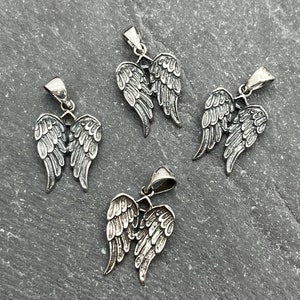 Op de afbeelding: Vier zilverkleurige engelenvleugelhangers. Elke hanger heeft gedetailleerde veren en een kleine lus voor een ketting. De hangers worden gepresenteerd op een getextureerd grijs oppervlak.