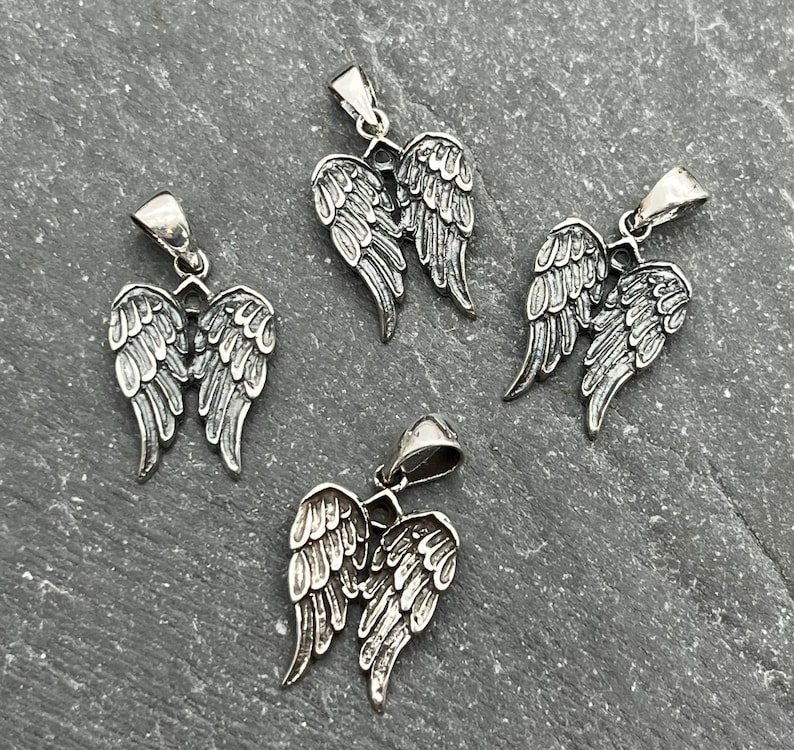 Op de afbeelding: Vier zilverkleurige engelenvleugelhangers. Elke hanger heeft gedetailleerde veren en een kleine lus voor een ketting. De hangers worden gepresenteerd op een getextureerd grijs oppervlak.