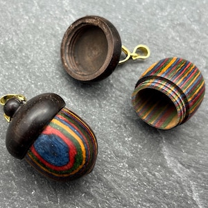 May include: Three wooden acorn-shaped pendants with a dark brown cap and a colorful striped body. One acorn is open, revealing the interior. The acorns have a gold-colored bail for attaching to a necklace or keychain.