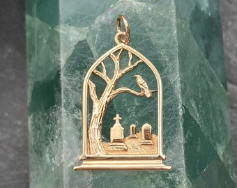 May include: Gold-colored pendant featuring a stylized graveyard scene. A tree with a perched bird is framed by an arched window, with tombstones in the foreground. The pendant is suspended from a small loop.