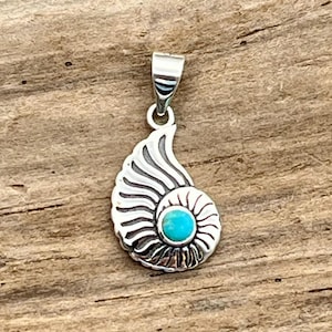 May include: A silver pendant in the shape of a nautilus shell. The shell design features detailed lines and a bright turquoise stone at the center. The pendant has a loop for a chain and is set against a wooden background.