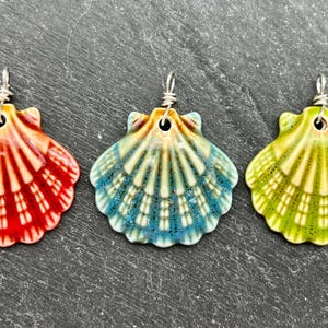 May include: Three seashell pendants in red, blue, and green. Each pendant is shell-shaped with a silver wire loop for hanging. The pendants have a glossy finish and are arranged on a dark gray surface.