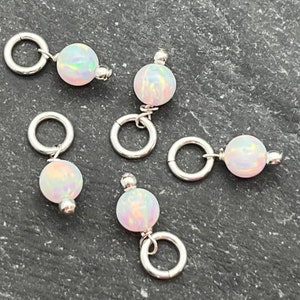 May include: Five small, iridescent opal beads with silver-colored metal rings and posts. The opal beads have a pearly white base color with flashes of blue, green, and pink. The metal rings are circular and attached to the beads.