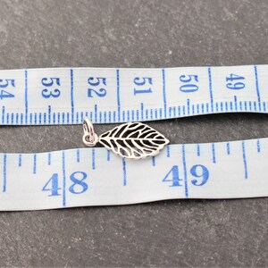Op de afbeelding: Een kleine, zilveren blad-hanger met gedetailleerde uitgesneden patronen. De hanger is geplaatst in de buurt van een meetlint voor schaal. Het ontwerp van de hanger is een bladvorm, met delicate aderdetails. Ongeveer 1,5 cm lang.