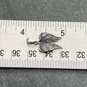 Op de afbeelding: Een zilverkleurige hanger in de vorm van engelen vleugels. De hanger is ongeveer 2,5 cm breed en 2 cm hoog. De vleugels zijn gedetailleerd met individuele veerrigels. De hanger ligt op een grijze ondergrond naast een liniaal.