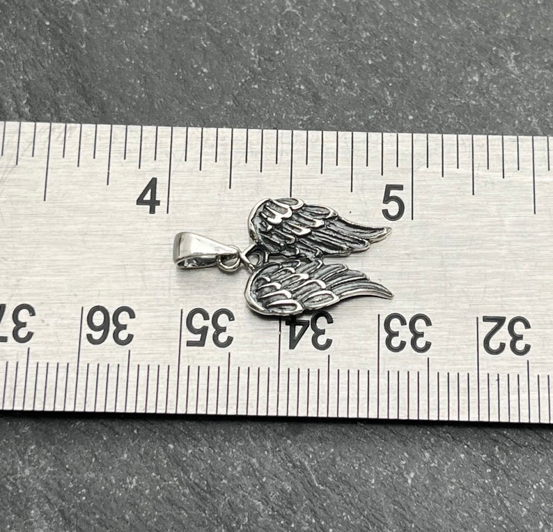 Op de afbeelding: Een zilverkleurige hanger in de vorm van engelen vleugels. De hanger is ongeveer 2,5 cm breed en 2 cm hoog. De vleugels zijn gedetailleerd met individuele veerrigels. De hanger ligt op een grijze ondergrond naast een liniaal.