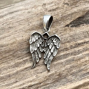 Op de afbeelding: Zilverkleurige hanger met een gedetailleerd engelenvleugelontwerp. De hanger heeft een lus aan de bovenkant voor een ketting en is geplaatst tegen een rustieke houten achtergrond. De vleugels zijn ingewikkeld gesneden met veerd details.
