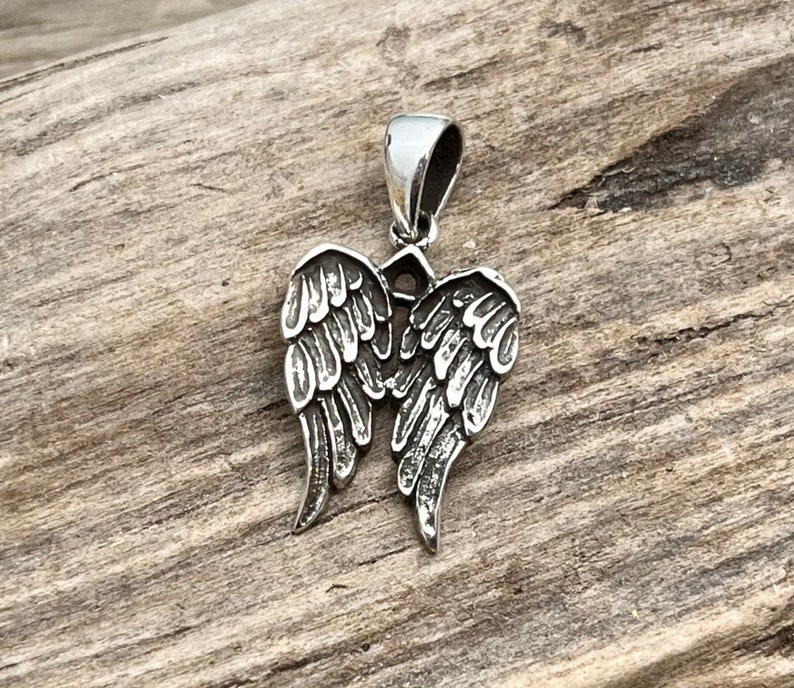 Op de afbeelding: Zilverkleurige hanger met een gedetailleerd engelenvleugelontwerp. De hanger heeft een lus aan de bovenkant voor een ketting en is geplaatst tegen een rustieke houten achtergrond. De vleugels zijn ingewikkeld gesneden met veerd details.