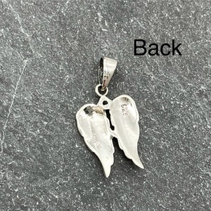 Op de afbeelding: Een zilveren hanger in de vorm van engelenvleugels. De hanger heeft een lus voor een ketting. Het woord "Back" is zichtbaar in de afbeelding. De hanger ligt op een donkergrijze ondergrond.