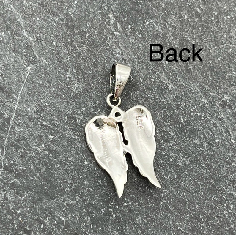 Op de afbeelding: Een zilveren hanger in de vorm van engelenvleugels. De hanger heeft een lus voor een ketting. Het woord "Back" is zichtbaar in de afbeelding. De hanger ligt op een donkergrijze ondergrond.