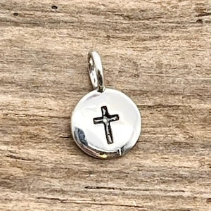May include: A small, round, silver-colored pendant with a cross design. The cross is centered on the pendant and has a simple, minimalist style. The pendant has a loop at the top for attaching to a chain or bracelet.