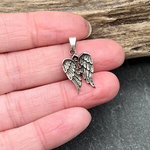 Op de afbeelding: Een kleine, zilverkleurige hanger met een gedetailleerd paar engelen vleugels. De vleugels zijn ingewikkeld ontworpen met veerdetaillering en zijn aan de bovenkant verbonden met een kleine lus voor een ketting. De hanger wordt getoond tegen een natuurlijke houten achtergrond.