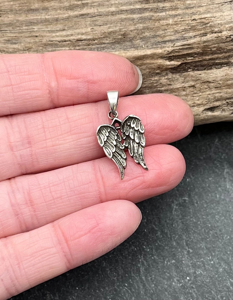 Op de afbeelding: Een kleine, zilverkleurige hanger met een gedetailleerd paar engelen vleugels. De vleugels zijn ingewikkeld ontworpen met veerdetaillering en zijn aan de bovenkant verbonden met een kleine lus voor een ketting. De hanger wordt getoond tegen een natuurlijke houten achtergrond.