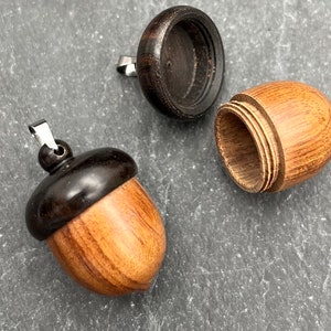 May include: A wooden acorn pendant, featuring a dark brown cap and a light brown body. The acorn is open, showing a hollow interior. A silver-coloured bail is attached to the cap for hanging. The acorn is made of wood.