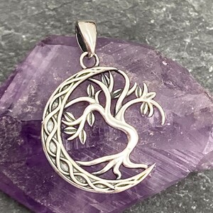 May include: Silver-toned pendant featuring a tree design within a crescent moon shape. The tree has detailed branches and leaves. The pendant has a bail for a chain. The background is a purple crystal.