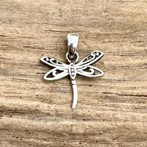 May include: A silver dragonfly pendant with intricate wing detailing. The dragonfly has a curved tail and a loop at the top for a chain. The pendant is set against a wooden background.