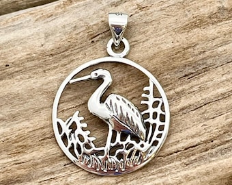 May include: A silver pendant featuring a detailed heron design within a circular frame. The heron is depicted in profile, standing amidst stylized reeds and grasses. The pendant has a bail for attaching to a chain.