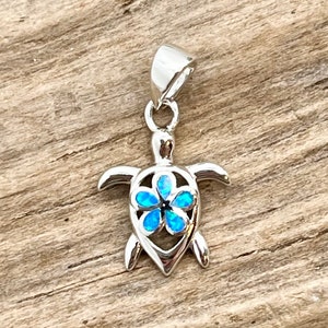 May include: A silver-colored pendant in the shape of a sea turtle. The turtle's shell features a blue flower design. The pendant has a loop at the top for a chain. The background is a wooden surface.