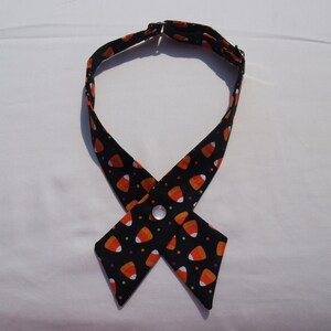 May include: A black Halloween-themed dog collar with a candy corn pattern. The collar is made of fabric and features a cross-over design with a button closure. The candy corn design includes orange, white, and yellow candy corn pieces on a black background.