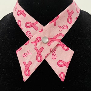 May include: A pink fabric necktie with a white button closure. The tie is patterned with pink ribbon designs.