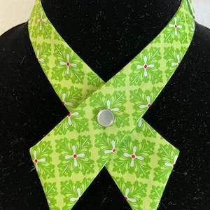 May include: A green and white patterned fabric collar with a silver button closure. The collar is designed to be worn as a neck accessory.