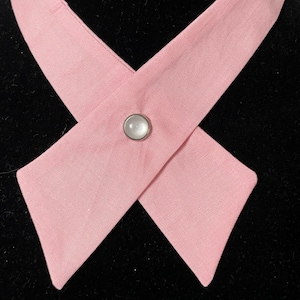 May include: A pink fabric collar with a white button in the center. The collar is crossed over and tied at the front.