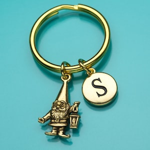 May include: Gold-toned keychain with a small gnome charm holding a lantern and a round charm with the letter "S" in black.