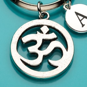 Puede incluir: Llavero plateado con un colgante con el símbolo Om y un charm circular con la letra "A". El símbolo Om es un diseño estilizado y circular. El llavero está sobre un anillo plateado, con un fondo turquesa.