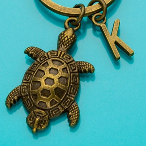 May include: A gold-toned turtle keychain with a letter K charm attached to a key ring. The turtle is detailed with a textured shell and has a three-dimensional design.