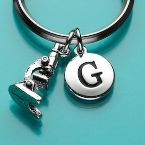 May include: Silver keychain with a microscope charm and a round charm with the letter G on it. The charms are attached to a silver key ring.