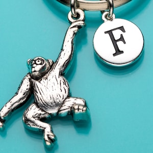 May include: Silver keychain with a monkey charm hanging from a silver ring with a silver charm with the letter F on it. The monkey charm is hanging by its arms and has a detailed face.
