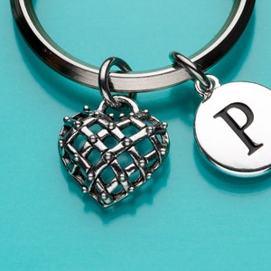 May include: Silver-toned keychain with a heart-shaped charm and a circular charm with the letter "P". The heart charm has a cage-like design. The keychain is on a turquoise background.