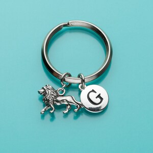 May include: Silver keychain with a lion charm and a round charm with the letter G. The keychain is on a turquoise background.