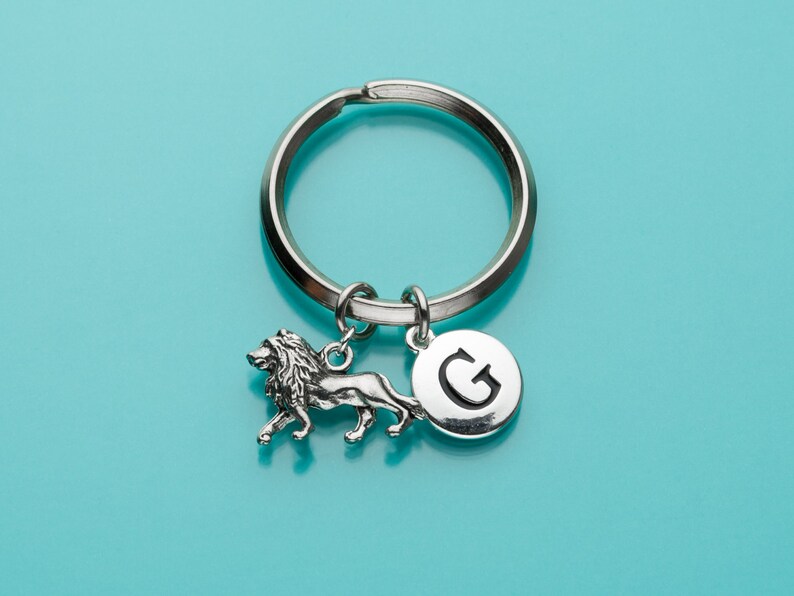 May include: Silver keychain with a lion charm and a round charm with the letter G. The keychain is on a turquoise background.