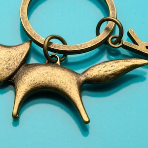 May include: A gold-toned metal keychain featuring a fox silhouette with a long tail. The keychain also has a metal ring and a letter 'K' charm.