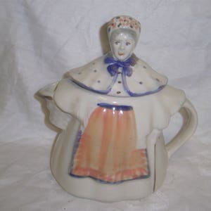 May include: A white ceramic teapot shaped like a woman wearing a blue and white dress with a pink apron. The teapot has a lid with a woman's head wearing a floral crown.